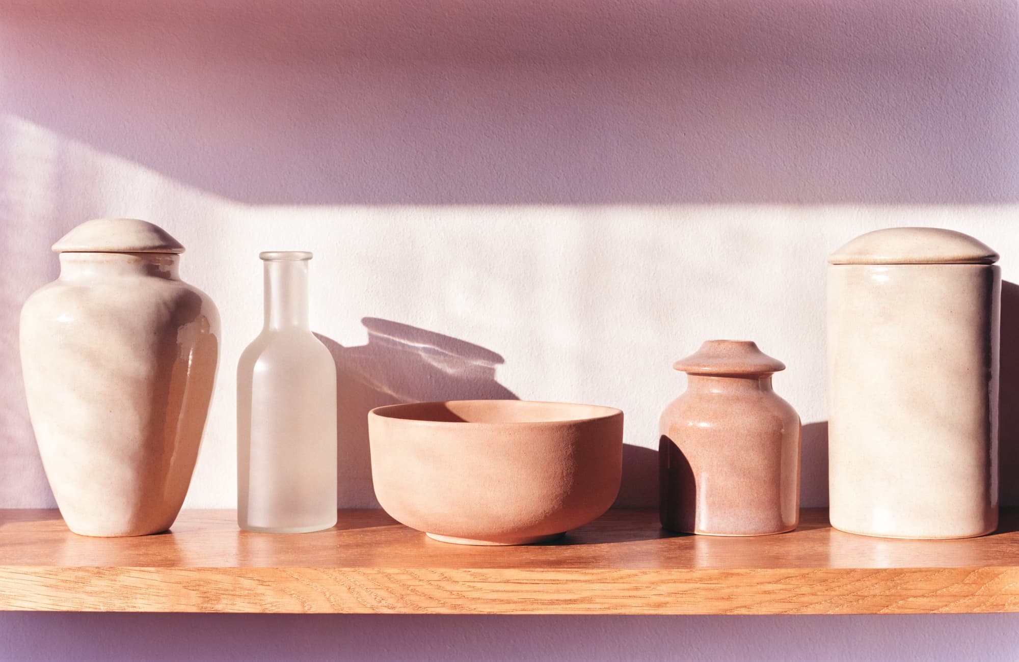 Five unlabeled ceramic vessels in ivory, clay and blush, evenly spaced on a warm oak shelf.