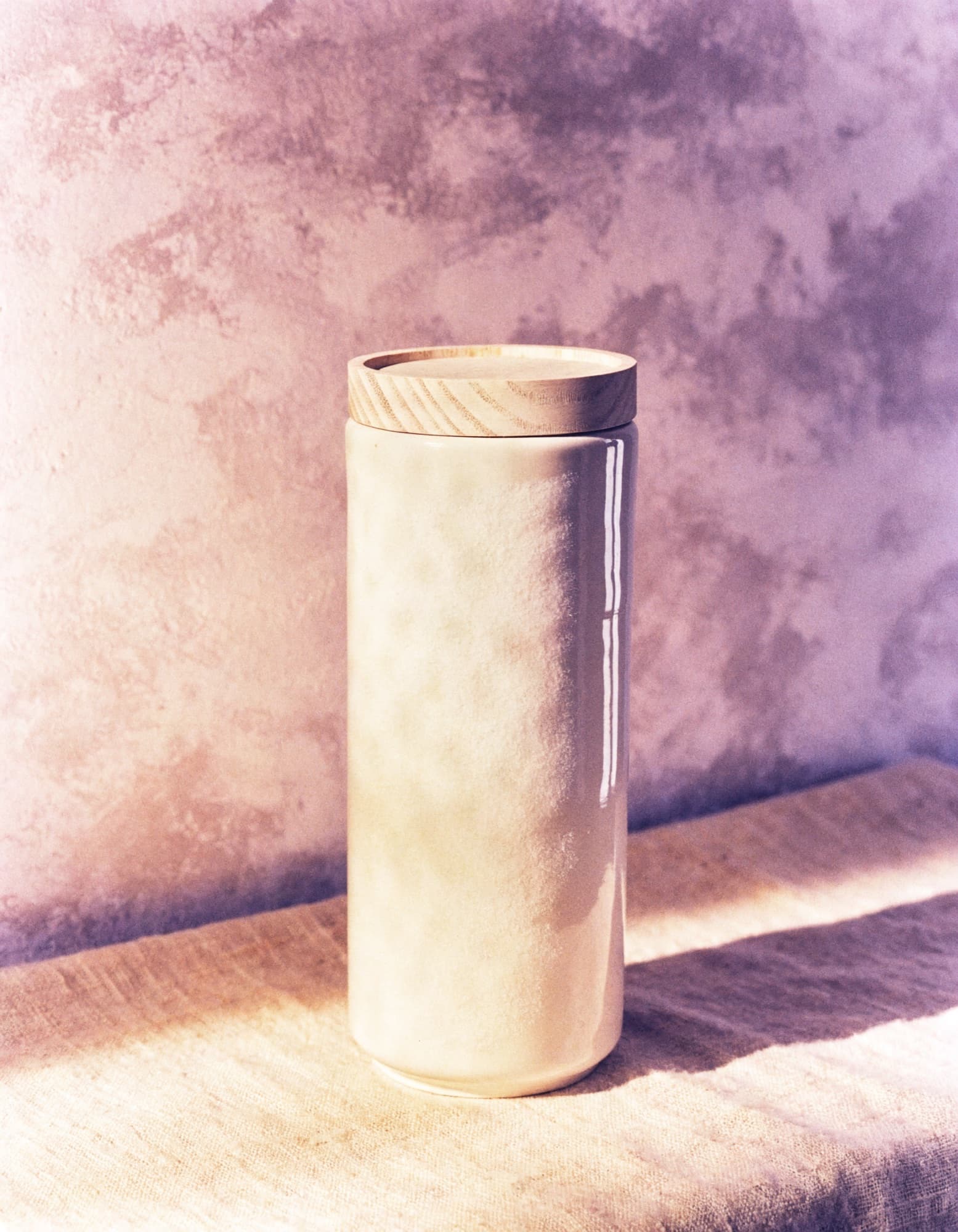 A tall unlabeled ivory ceramic jar with a pale wooden lid, side-lit against a dusty lavender plaster wall.