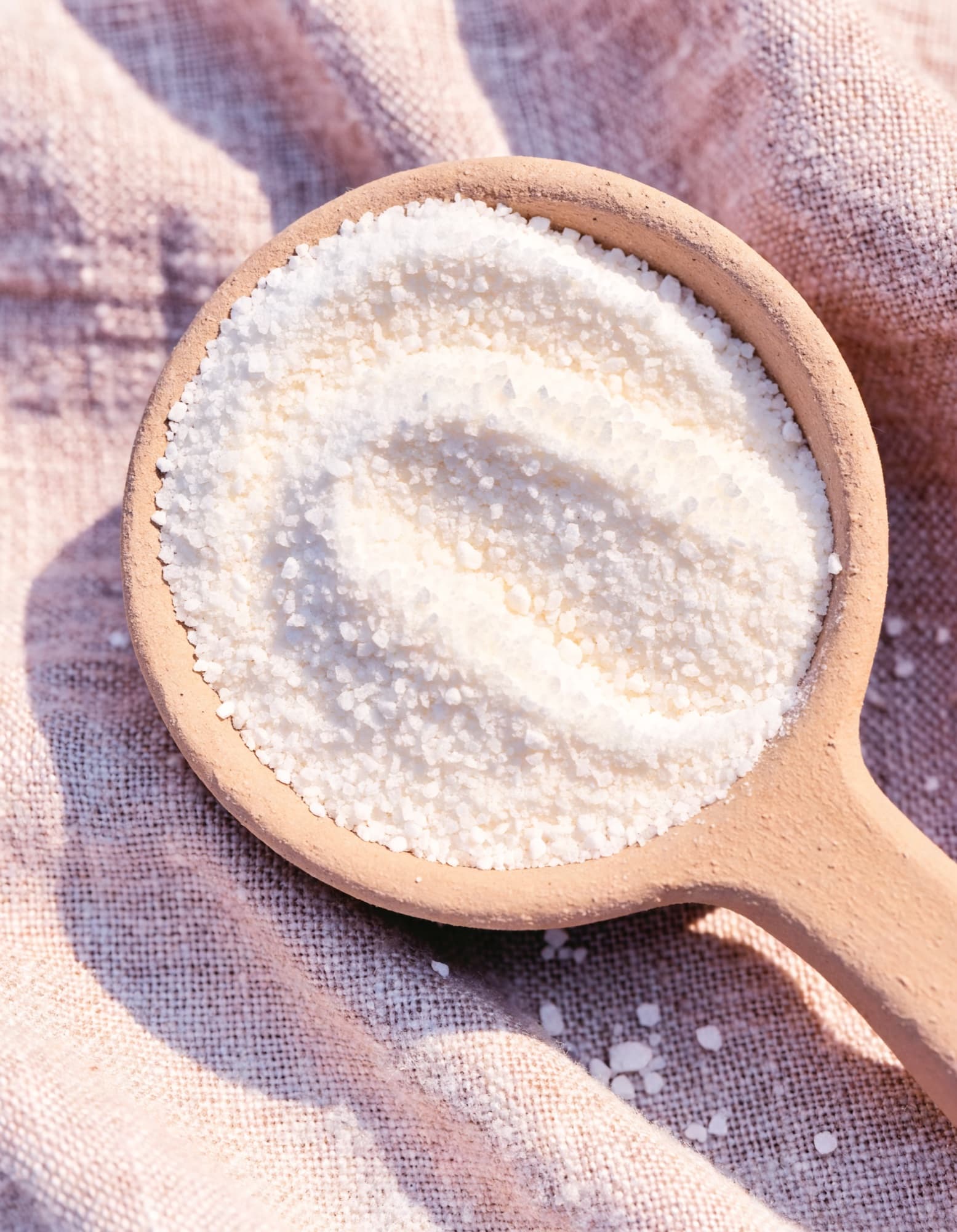 A wooden-handled scoop heaped with fine white laundry powder, resting on blush and lavender linen.
