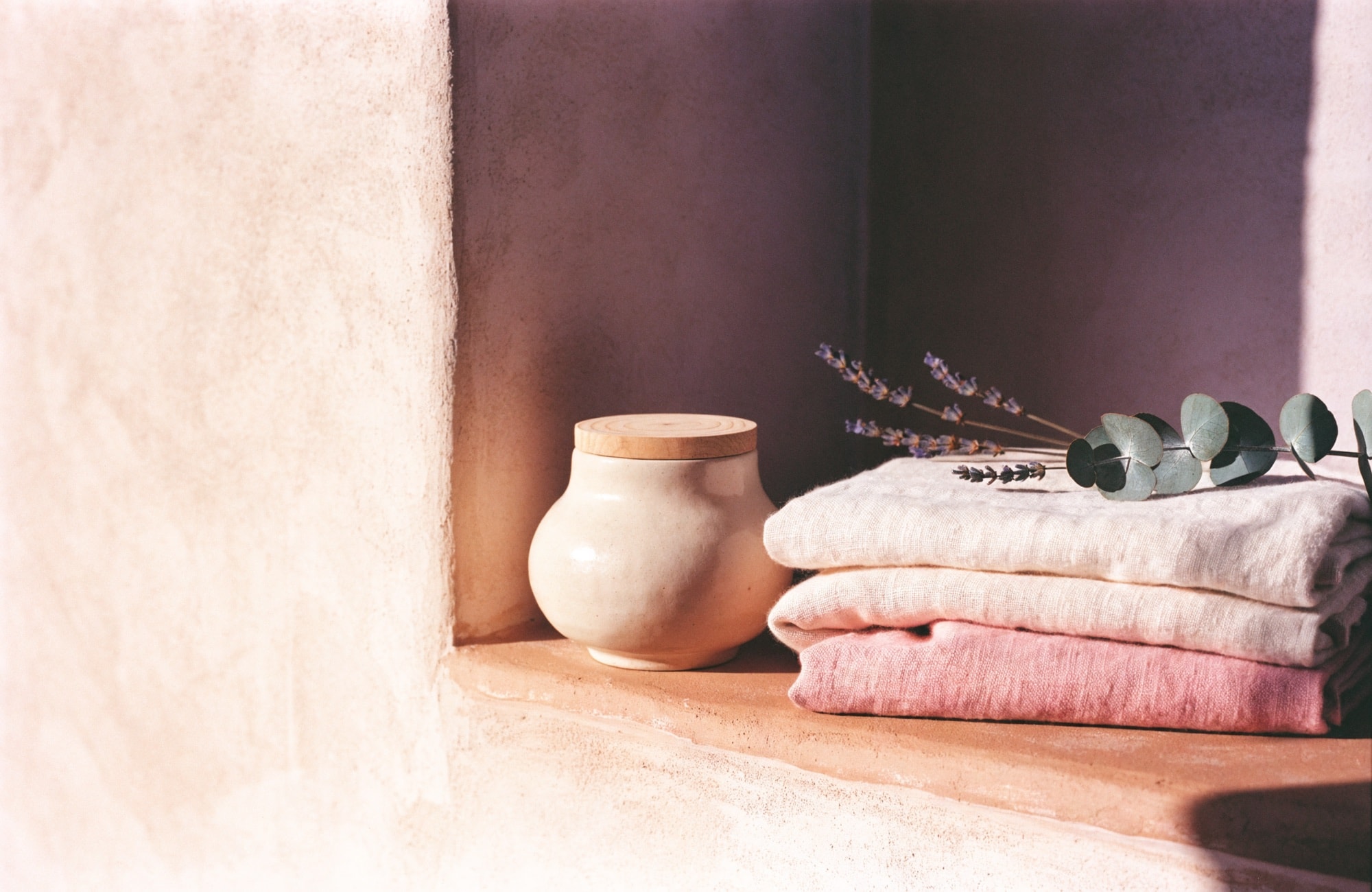An unlabeled ivory vessel on a warm plaster ledge beside folded blush linens and dried lavender.